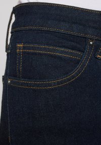 Jeans de mezclilla azul oscuro con costura amarilla, que cuentan con un bolsillo trasero y lazos para cinturón. La tela tiene una textura suave y un ligero brillo.