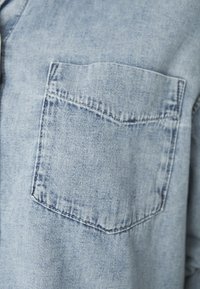 Camisa de ganga azul claro com um bolso no peito. O tecido tem uma textura suave e os detalhes de costura delineiam as bordas do bolso.