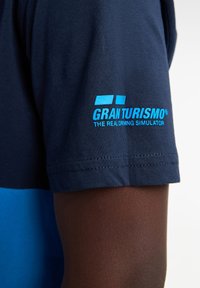 Námořnicky modré tričko s krátkým rukávem, na levém rukávu s modrým logem „GRAN TURISMO® THE REAL DRIVING SIMULATOR“.}}
