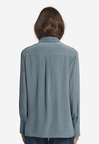 Kvinna med axellångt brunt hår, iklädd en långärmad grå blus och mörkblå jeans, sedd bakifrån mot en vit bakgrund.