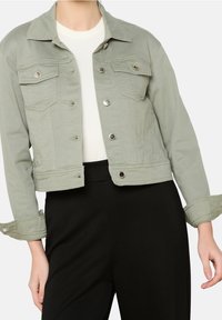 Osoba ubrana w oliwkową denimową kurtkę na białej bluzce i czarnych spodniach, z podwiniętymi rękawami i niezapiętą kurtką.