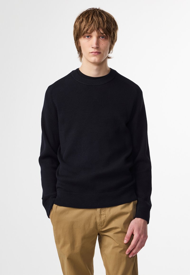 Svart stickad tröja med ribbad textur och rundade halsringning. Matchas med beige byxor, som har en avslappnad passform och rak ben-design.