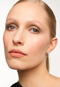 Primo piano di un volto con pelle liscia, rossetto nude opaco, occhi marroni chiari e ombretto dorato. I capelli sono lisci e raccolti. Orecchini a cerchio in argento.