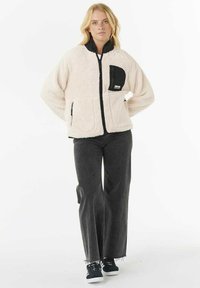 Blonde vrouw staat, gekleed in een crèmekleurige fleecejack met zwarte rand en zakken, donkere wijde jeans en zwarte sneakers.