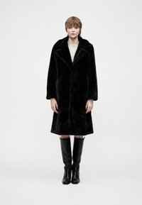 Pessoa em pé, de frente, vestindo um casaco de pelúcia preta, uma blusa bege e botas de couro pretas até ao joelho, em frente a um fundo branco liso.