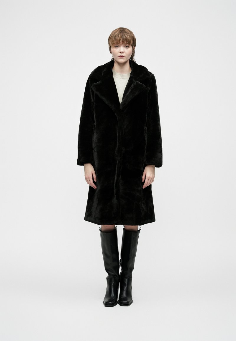 Pessoa em pé, de frente, vestindo um casaco de pelúcia preta, uma blusa bege e botas de couro pretas até ao joelho, em frente a um fundo branco liso.