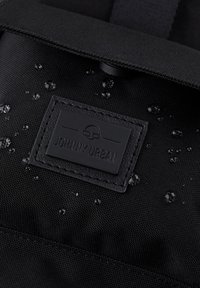 Rucsac negru rezistent la apă, cu un material texturat, având o etichetă din piele branduită și picături pe suprafață, indicând protecție împotriva umezelii.