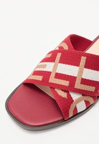 Sandale rouge avec des brides croisées en tissu tressé, présentant un motif géométrique en beige et blanc. Semelle plate au toucher lisse.