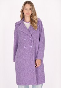 Cappotto doppio petto viola, soffice, con colletto a punte, caratterizzato da bottoni rotondi viola e maniche lunghe, indossato sopra dei jeans chiari.