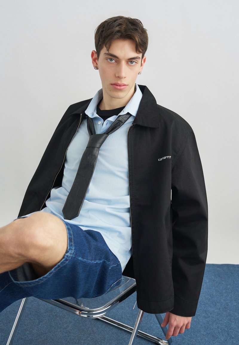 Νέος άνδρας καθισμένος σε καρέκλα φορώντας μαύρο σακάκι, ανοιχτό μπλε πουκάμισο, μαύρη γραβάτα, τζιν σορτς και σκουλαρίκια μπροστά σε μονόχρωμο φόντο.