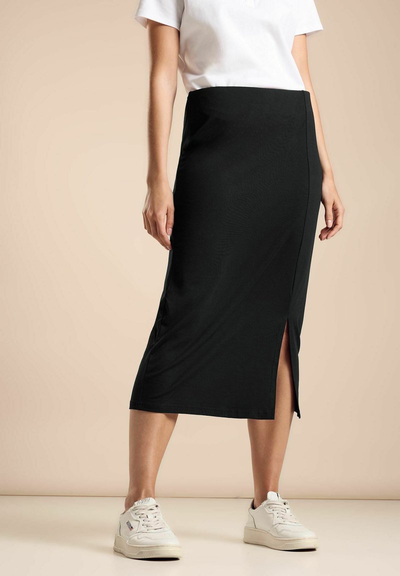Gonna nera midi con uno spacco laterale, realizzata in tessuto morbido. Indossata con una t-shirt bianca e sneakers chiare, in piedi su uno sfondo chiaro.
