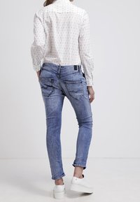 Jean skinny en denim bleu avec une finition délavée, comportant deux poches arrière et des détails de couture, associés à une chemise blanche à motifs et des chaussures blanches.