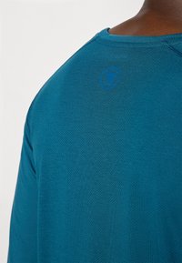 Teal Langarmshirt aus einem strukturierten, atmungsaktiven Material; besitzt ein kleines blaues Logo am Nacken hinten und kontrastierende Nähte.