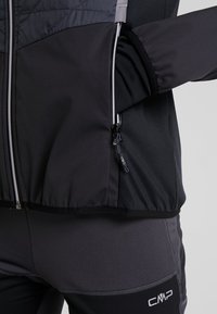 Personne portant une veste de sport CMP noire et grise avec un pantalon, la main placée à l'intérieur de la fermeture éclair de la poche de la veste.