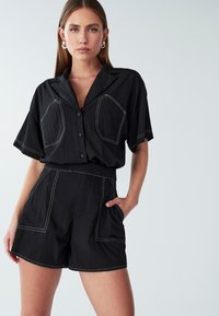 Mladá žena oblečená v čiernej košeli s krátkym rukávom na gombíky a zodpovedajúcich šortkách s vysokým pásom, ktoré majú biele stehy, pózuje s jednou rukou v vrecku.