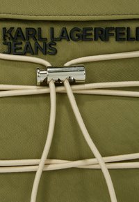 Grüner Stoff mit strukturiertem Finish, mit schwarzem Logo "KARL LAGERFELD JEANS" und metallischen Beschlägen, durch die beiges elastisches Band gewoben ist.