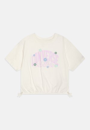Camiseta corta blanca de manga corta con texto "Converse All Star" en estampados florales en tonos pastel púrpura y azul, con cordones ajustables en el dobladillo.