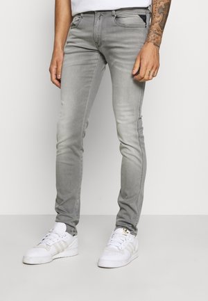 Homme portant un jean gris slim-fit et des baskets blanches, debout devant un fond clair uni, avant-bras droit tatoué visible.