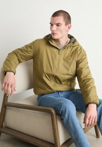 Olivegrüne, leichte Jacke aus Nylon, mit Reißverschluss vorne, verstellbarer Kapuze und elastischen Bündchen, kombiniert mit hellblauen Jeans.