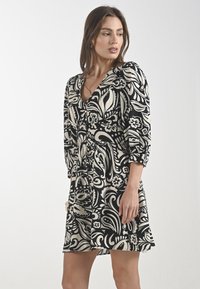 Una donna in un vestito nero e beige a fantasia con scollo a V e maniche a tre quarti si trova davanti a uno sfondo neutro, guardando di lato.