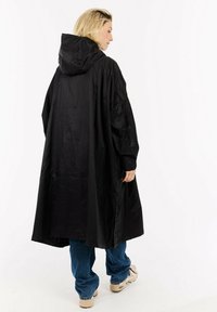 Zwarte waterdichte lange jas met een capuchon, met een losse pasvorm en een zachte textuur. Draagt over blauwe spijkerbroeken en lichtgekleurde schoeisel.