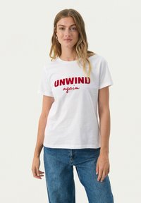 Witte katoenen shirt met rode tekst die "UNWIND" en "again" in een casual lettertype leest. Korte mouwen en standaard pasvorm.