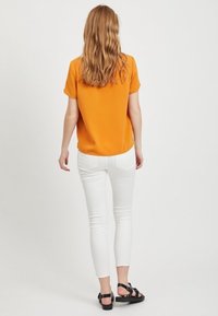 Orange short-sleeve top, white skinny jeans, and black sandals. The top has a rounded neckline; the jeans are slim-fitting with a smooth texture.