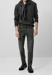 Maglione grigio con cappuccio, indossato sopra una camicia bianca. Jeans grigio scuro con tasche frontali e stivali alla caviglia neri. Design minimalista.