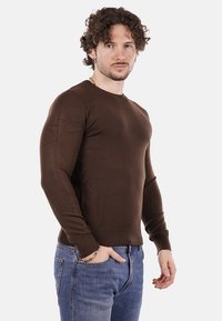 Uomo con i capelli ricci che indossa una camicia a maniche lunghe marrone e jeans blu, in piedi con una mano in tasca contro uno sfondo bianco.