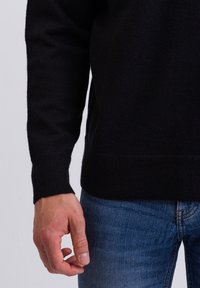 Schwarzer Strickpullover mit gerippten Bündchen und Saum, kombiniert mit blauen Jeans, wobei eine Hand auf Hüfthöhe positioniert ist.