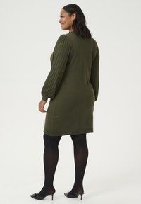 Robe tricotée vert olive avec des manches bouffantes côtelées, longueur genoux, associée à des collants noirs et des chaussures à talons. Ourlet simple et silhouette ajustée.