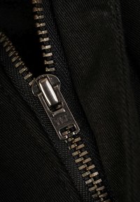 Tela negra con un primer plano de una cremallera metálica YKK, con dientes suaves y una lengüeta rectangular. Fondo texturizado visible.