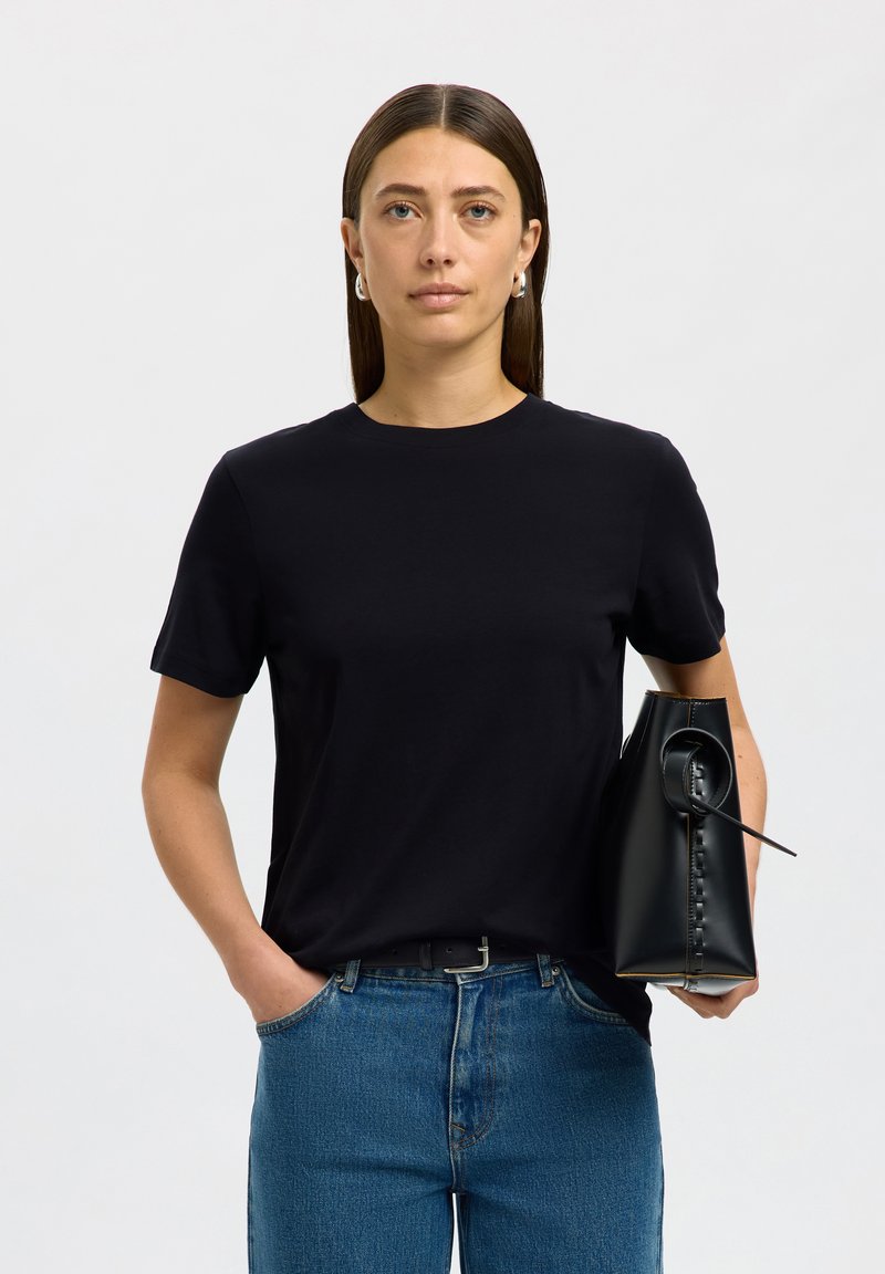 Frau mit glatten braunen Haaren, die ein schwarzes T-Shirt und blaue Jeans trägt, hält eine schwarze Ledertasche und steht vor einem einfarbigen Hintergrund.