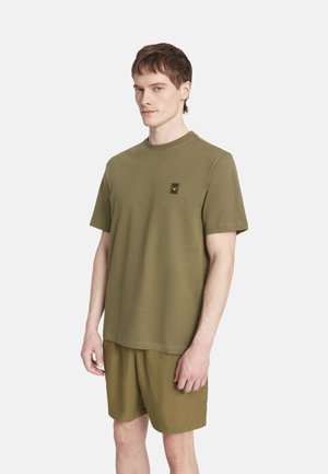 Uomo che indossa una t-shirt verde oliva a maniche corte con piccolo logo sul petto e pantaloncini coordinati su uno sfondo chiaro e uniforme.