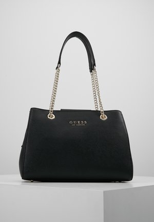 Sac à main Guess noir avec anses en chaîne dorée et logo "GUESS Los Angeles" centré sur le devant, exposé sur un piédestal blanc.
