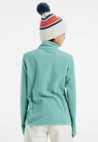 Giacca in pile turchese con colletto alto, abbinata a un berretto di maglia bianco con pom-pom blu e strisce rosso-blu.