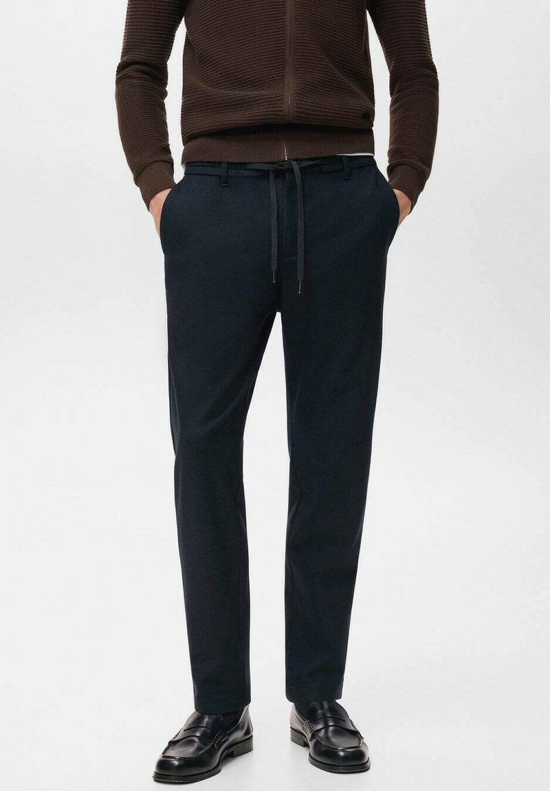 Pantalon bleu marine avec une coupe ajustée, fabriqué en tissu doux. Comprend un cordon de serrage et des poches latérales. Associé à des chaussures en cuir noir.