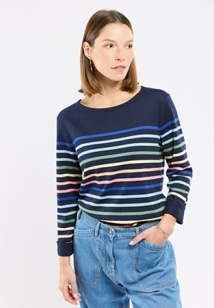Femme portant un pull bleu marine à rayures horizontales multicolores et un jean en denim taille haute bleu clair, posant avec une main dans la poche.