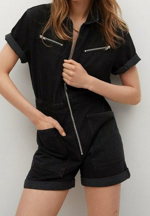 Femme portant une combinaison courte en jean noire à manches courtes avec fermeture éclair devant, manches retroussées, poches poitrine zippées et grandes poches plaquées devant.