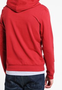 Sudadera de algodón roja con un bolsillo frontal y capucha con cordón, con puños y dobladillo acanalados, combinada con jeans oscuros.