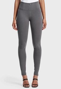 Nainen, jolla on päällään korkeavyötäröiset, tummanharmaat, etuvetoketjulliset kapeat housut, yhdistettyinä mustiin avojalkaisiin korkokenkiin vaaleaa taustaa vasten.