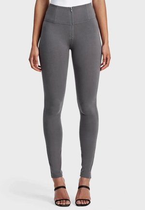 Nainen, jolla on päällään korkeavyötäröiset, tummanharmaat, etuvetoketjulliset kapeat housut, yhdistettyinä mustiin avojalkaisiin korkokenkiin vaaleaa taustaa vasten.