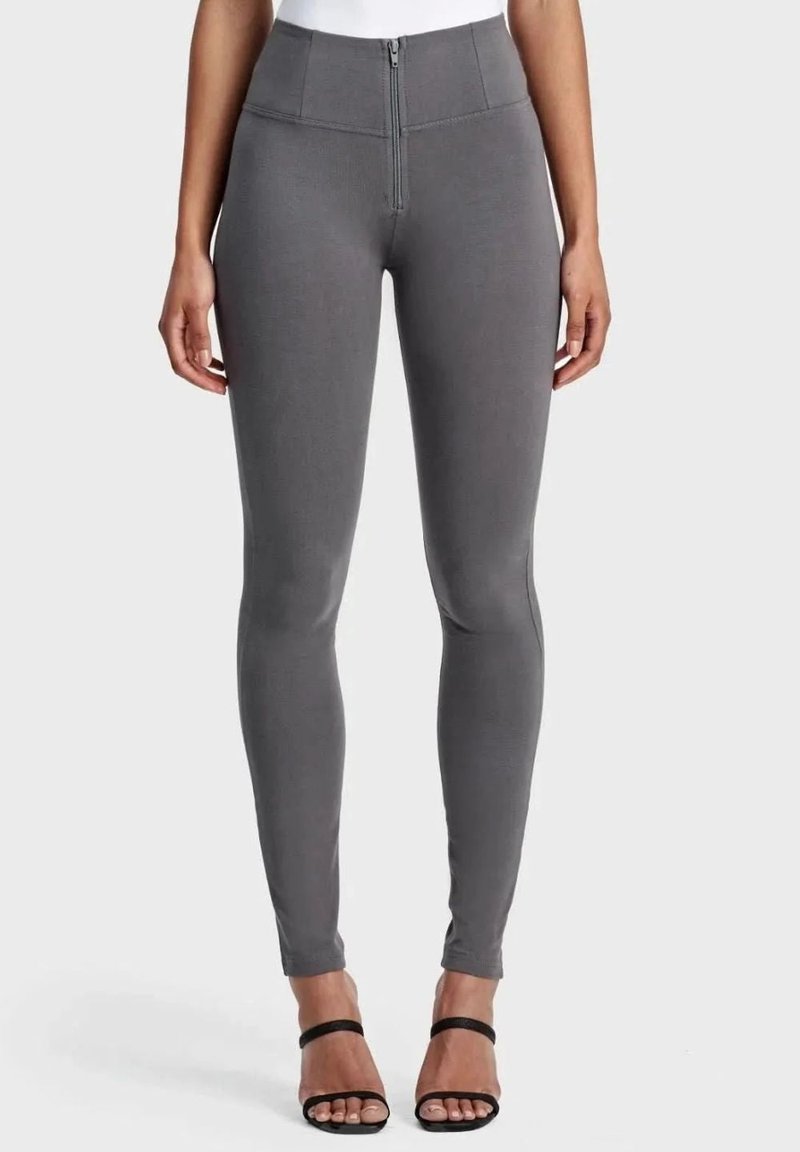 Nainen, jolla on päällään korkeavyötäröiset, tummanharmaat, etuvetoketjulliset kapeat housut, yhdistettyinä mustiin avojalkaisiin korkokenkiin vaaleaa taustaa vasten.