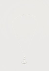 Λεπτό ασημένιο κολιέ με αλυσίδα και μικρό μενταγιόν σε σχήμα άγκυρας, σε απλό φωτεινό φόντο.