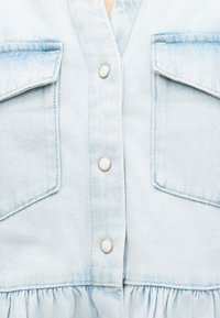 Φόρεμα από ανοιχτό μπλε τζιν με δύο τσέπες με καπάκι και μεταλλικά κουμπιά με πατούρα κατά μήκος της κεντρικής ραφής.