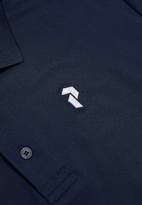 Navy polo met een gestructureerde afwerking, voorzien van een witte geborduurde logo en een knopenlijst. Inclusief een kraag en korte mouwen.