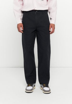 Man wearing black wide-leg pants, white sneakers with black and yellow details, and a light pink button-up shirt standing against a white background.