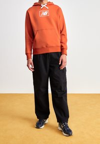 Felpa con cappuccio arancione, logo bianco e tasca frontale, abbinata a pantaloni cargo neri e scarpe da ginnastica blu. Design semplice, vestibilità casual.