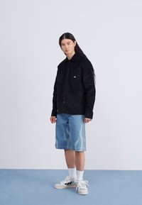 Camisa negra de botones con un bolsillo en el pecho, combinada con pantalones cortos de mezclilla azul claro con patrones de llamas. Calcetines deportivos blancos y zapatillas.