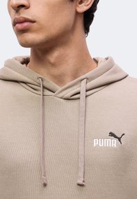 Νέος με μπεζ φούτερ Puma με ορατό λογότυπο, που δείχνει τον λαιμό και το κάτω μέρος του προσώπου σε ένα καθαρό φωτεινό φόντο.